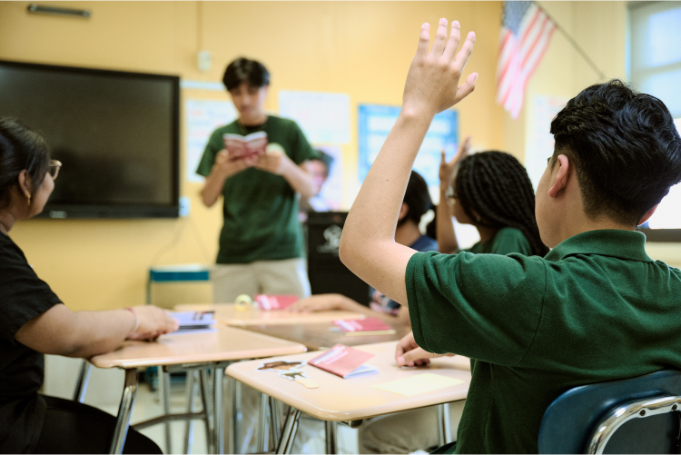 Students wearing green uniforms in a classroom. One student is raising a hand while another reads aloud. An American flag is visible in the background.